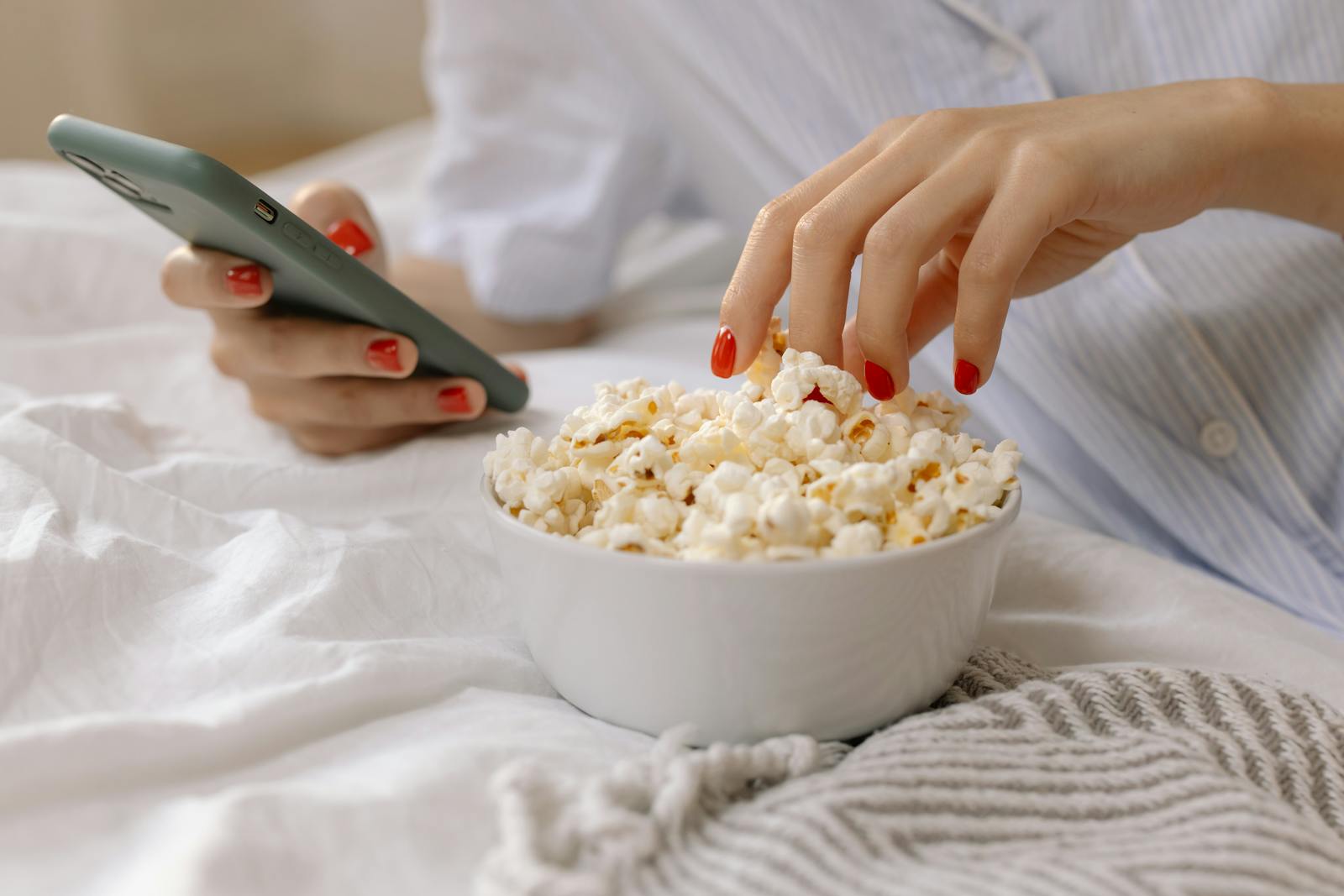 Close-up of a woman's hand with red nails using a smartphone while eating popcorn on bed.